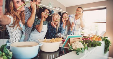 Friends wearing asparagus stalks under their noses while standing in front of table full of pots, pasta noodles and vegetables (Friends wearing asparagus stalks under their noses while standing in front of table full of pots, pasta noodles and vegetab